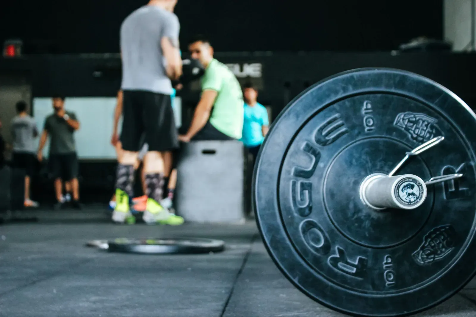A loaded barbell on a gym floor with athletes training behind it